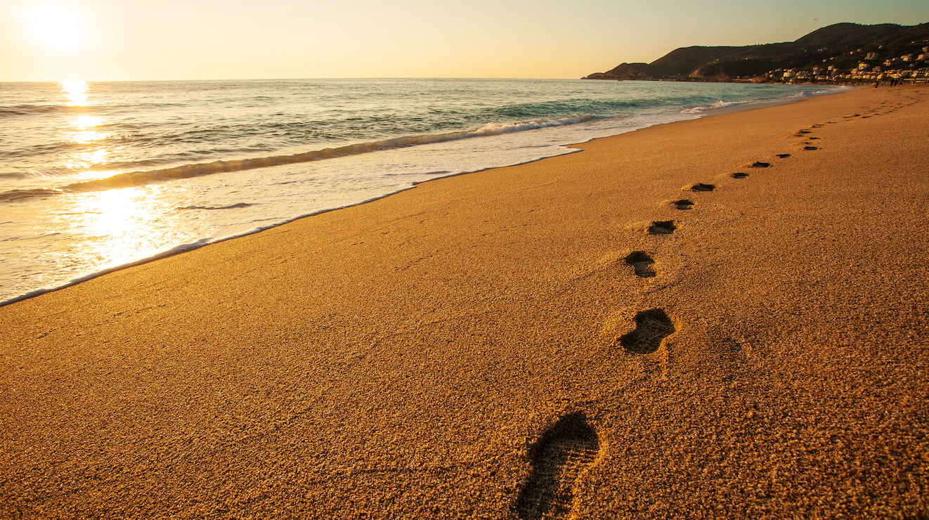 Le impronte sulla sabbia bagnata conducono lungo una spiaggia tranquilla al tramonto, con le onde che lambiscono dolcemente la riva: ogni passo è simbolo di ricordi ed eredità, come un lascito testamentario sotto un cielo dorato e colline in lontananza.