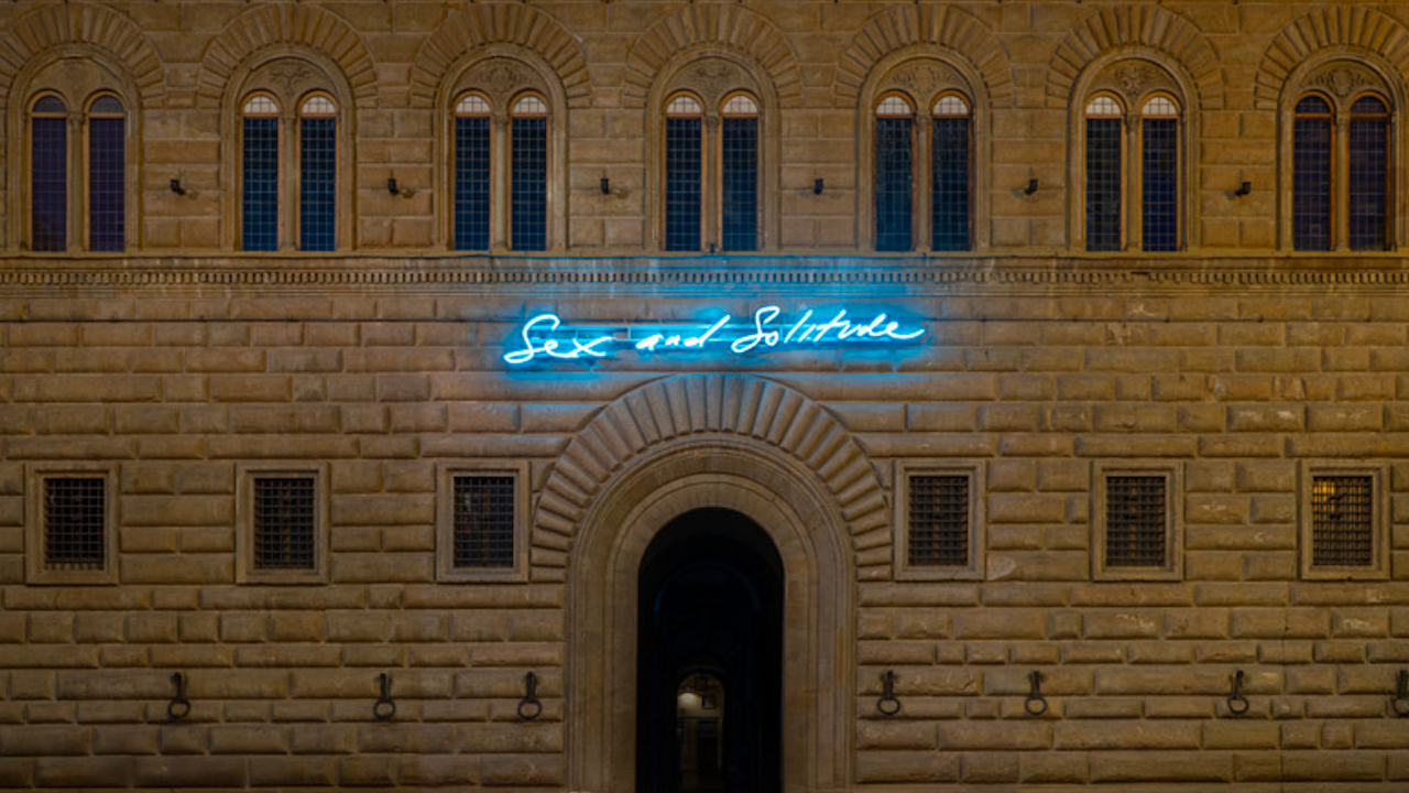 Un edificio in pietra con finestre ad arco è illuminato da un'insegna al neon blu sopra l'ingresso, su cui è scritto in corsivo "Sesso e solitudine".