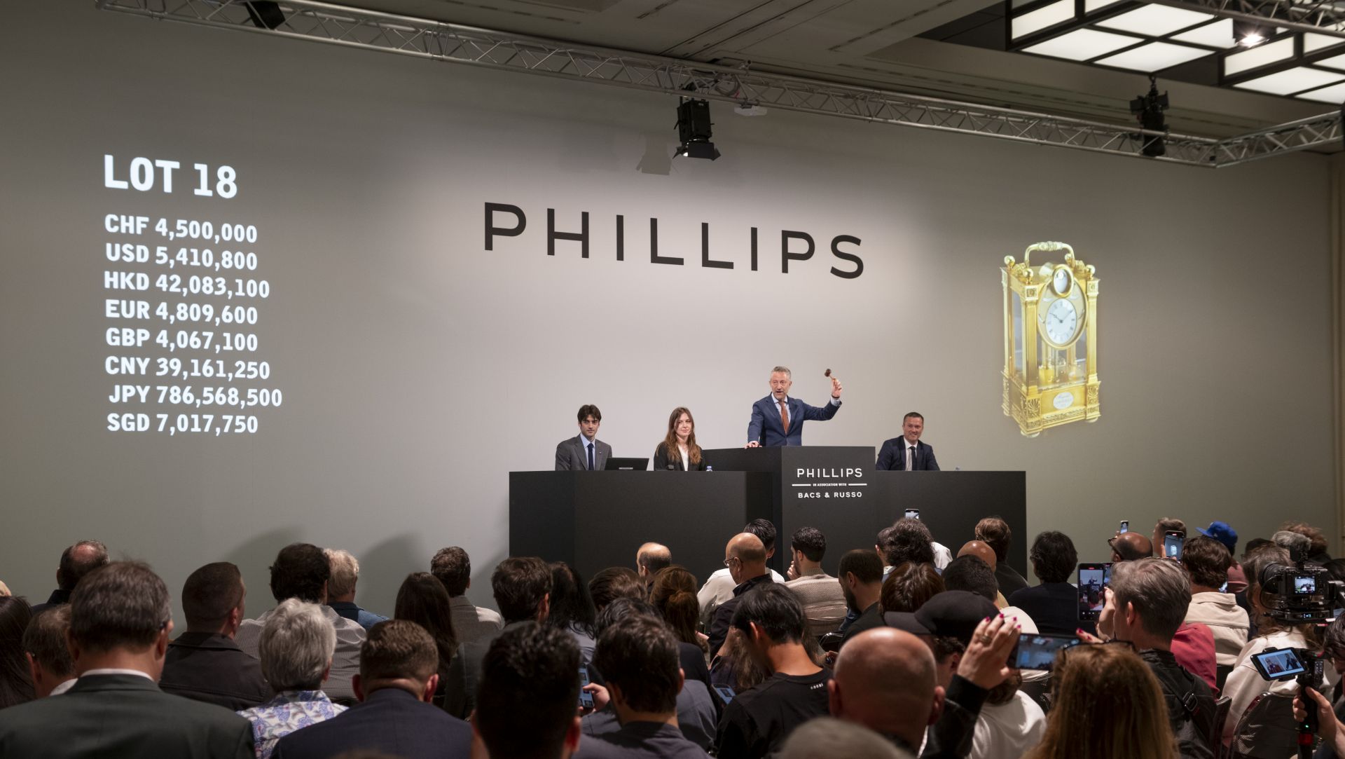 Un banditore d'asta è in piedi su un podio durante un'asta affollata da Phillips, con un grande orologio esposto alla parete e diversi valori di valuta elencati per il Lotto 18 su uno schermo. Diverse persone sono sul palco e il pubblico scatta foto.