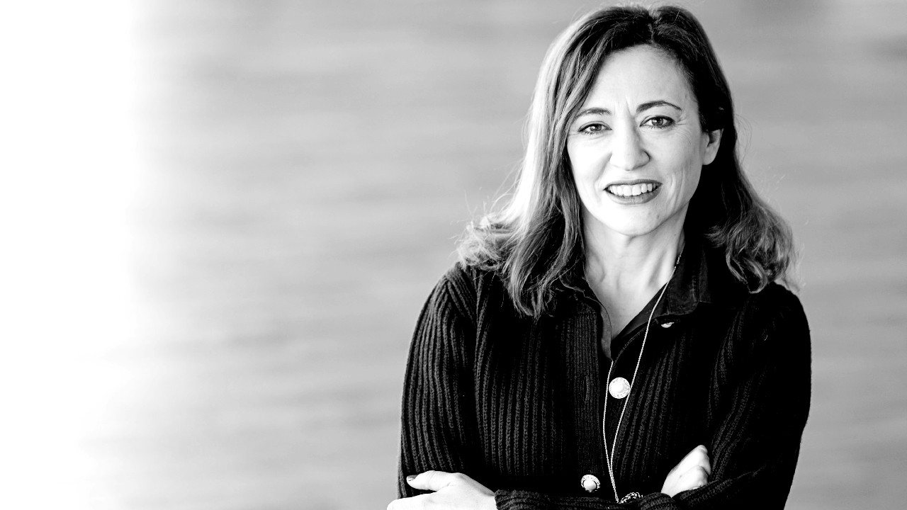 In questa foto in bianco e nero, una donna con i capelli mossi, che indossa un maglione scuro e una collana, è in piedi, sorridente, con le braccia incrociate su uno sfondo sfocato.