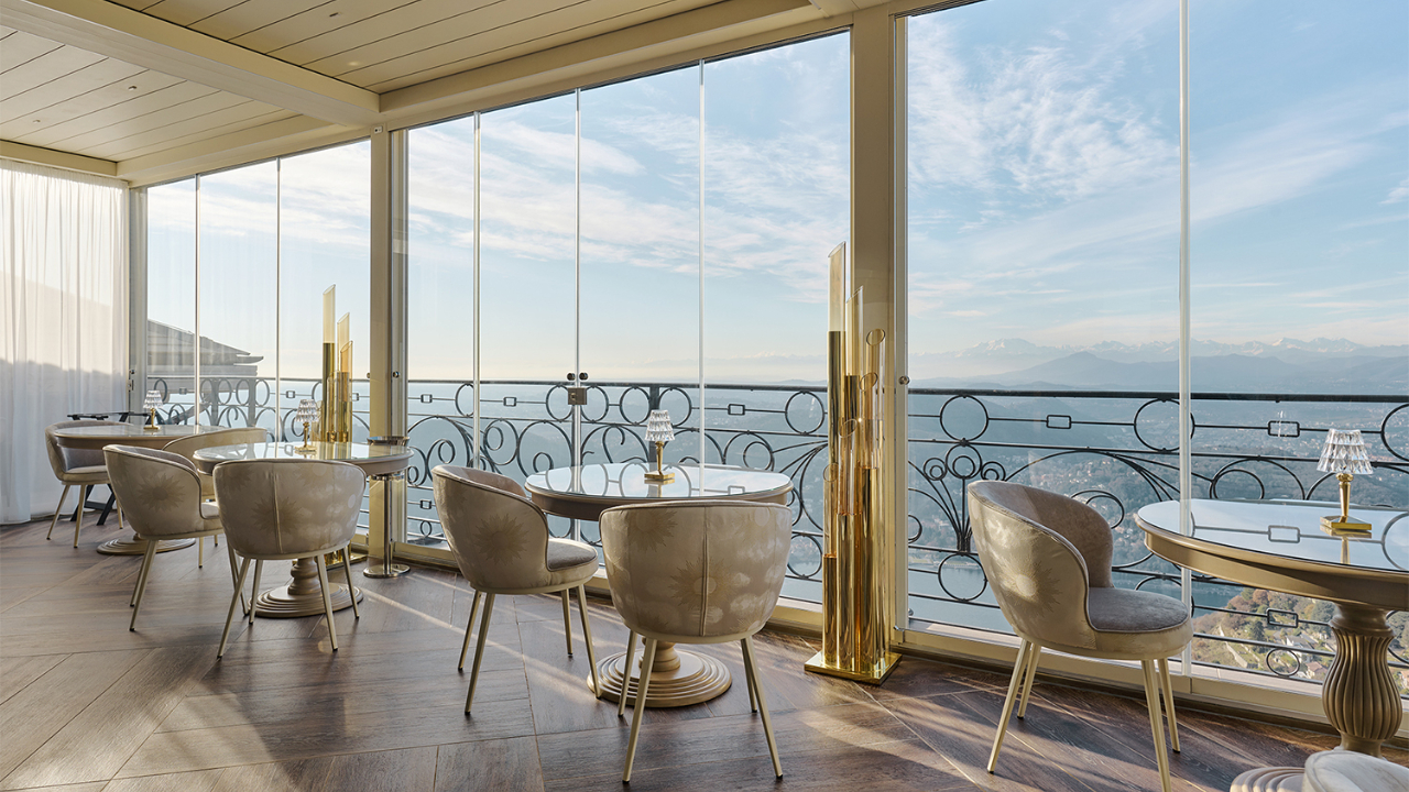Un ristorante moderno con tavoli e sedie eleganti, finestre a tutta altezza e una vista panoramica sulle montagne e sulla valle sotto un cielo azzurro con nuvole, proprio come quelle che si vedono a Como.