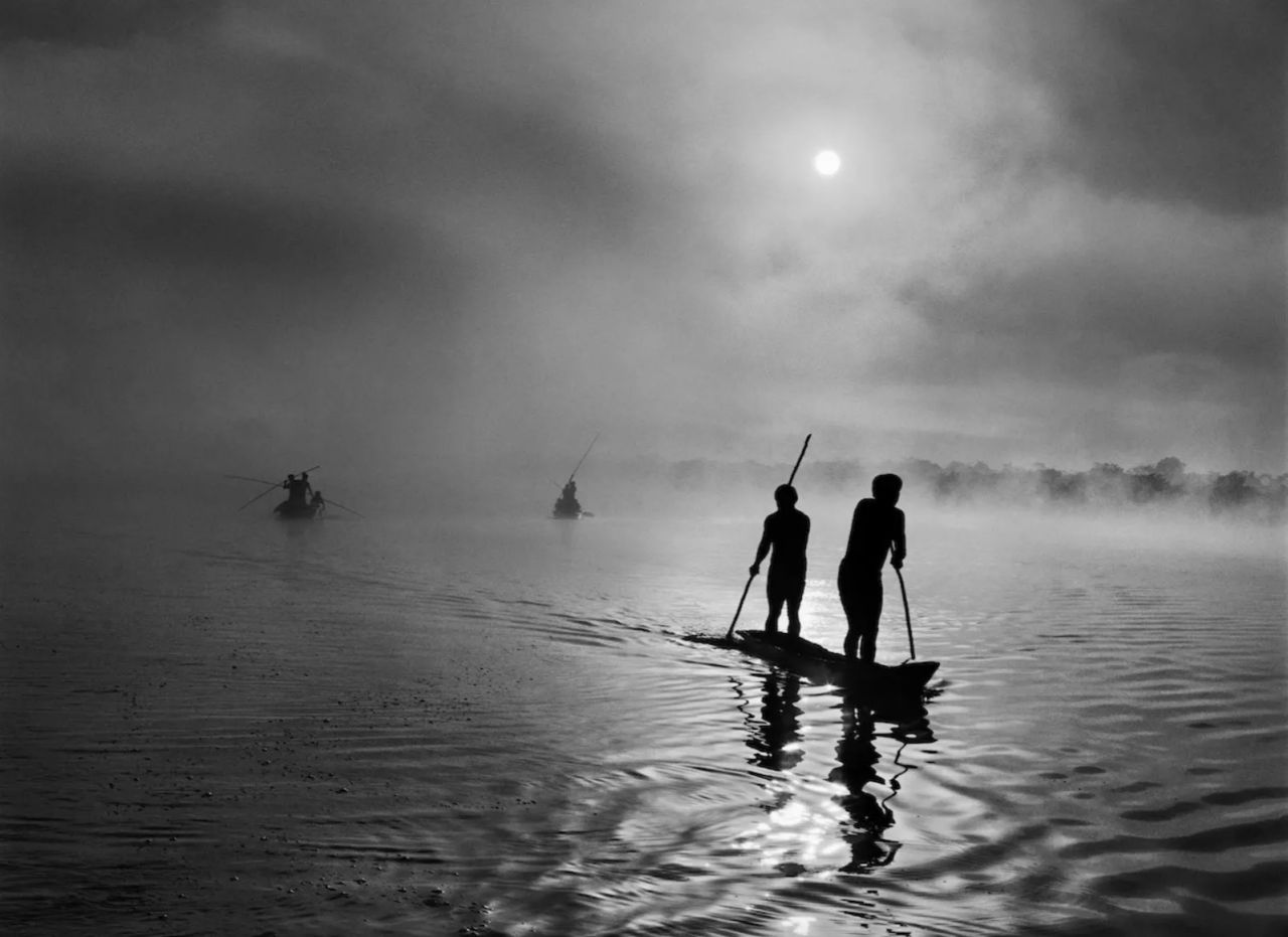 Sagome di due persone che remano in canoa su un fiume nebbioso all'alba, con altre barche in lontananza e la nebbia che ricopre l'acqua e gli alberi sullo sfondo.