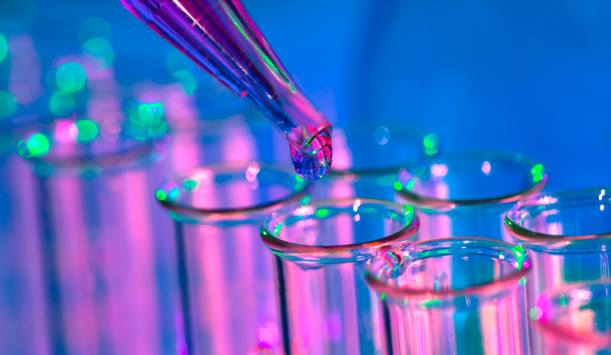 A close-up of a pipette dispensing a drop of liquid into one of several clear test tubes, illuminated with vibrant pink, blue, and green lighting in a laboratory setting.
