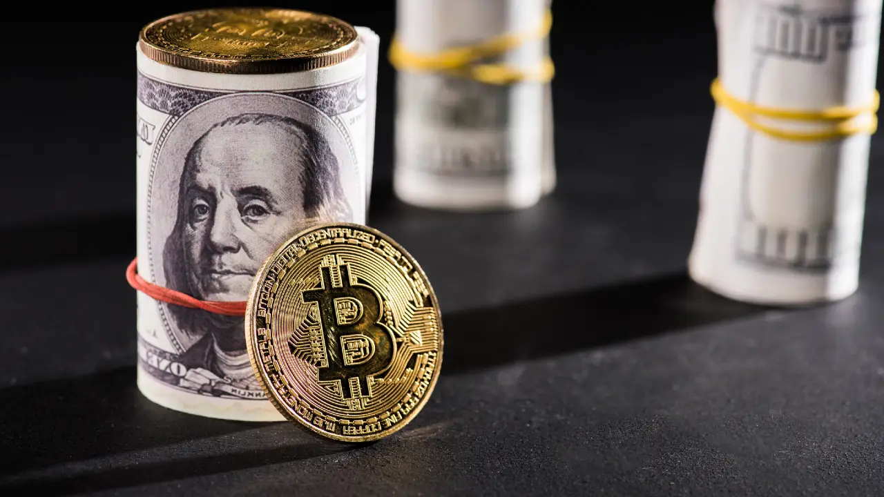 Una moneta Bitcoin d'oro è appoggiata su una banconota da cento dollari arrotolata e fissata con un elastico rosso, con altre banconote arrotolate sullo sfondo sfocato su una superficie scura.