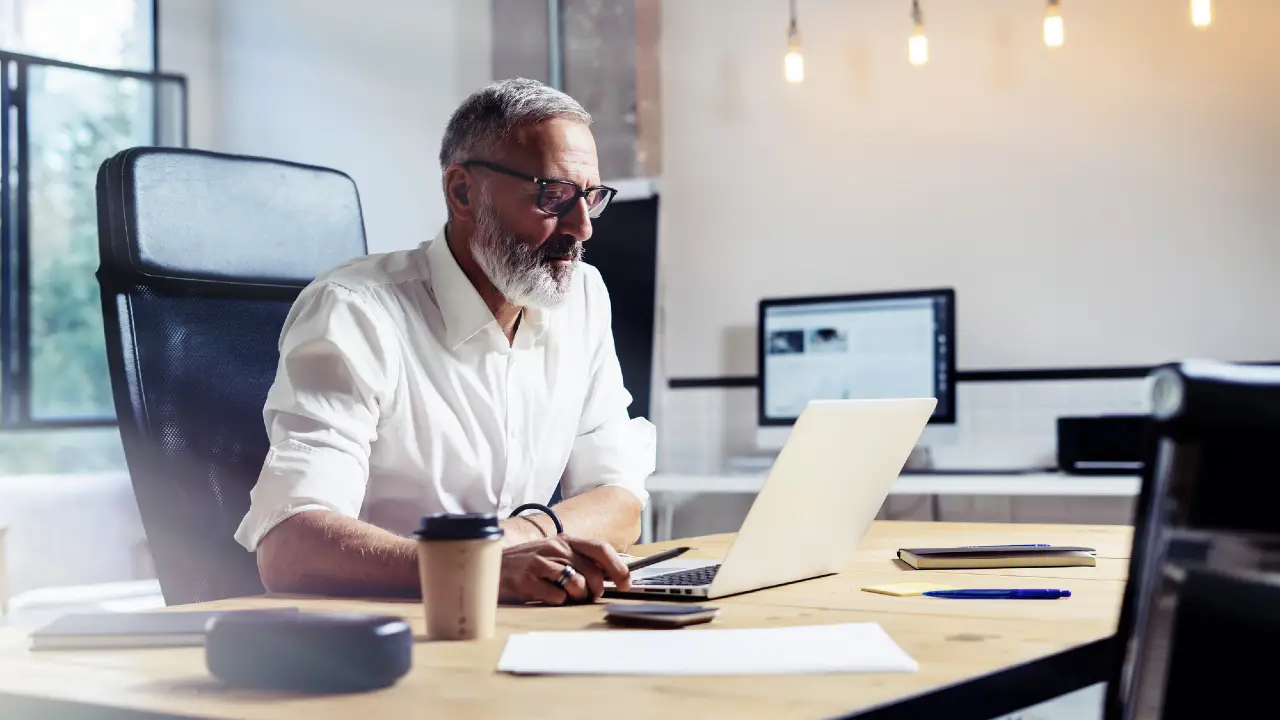 Un uomo con i capelli grigi e la barba, occhiali e camicia bianca, è seduto a una scrivania e lavora al computer portatile in un ufficio moderno. Sulla scrivania ci sono una tazza di caffè, un quaderno e una penna. Sullo sfondo si vedono schermi di computer.