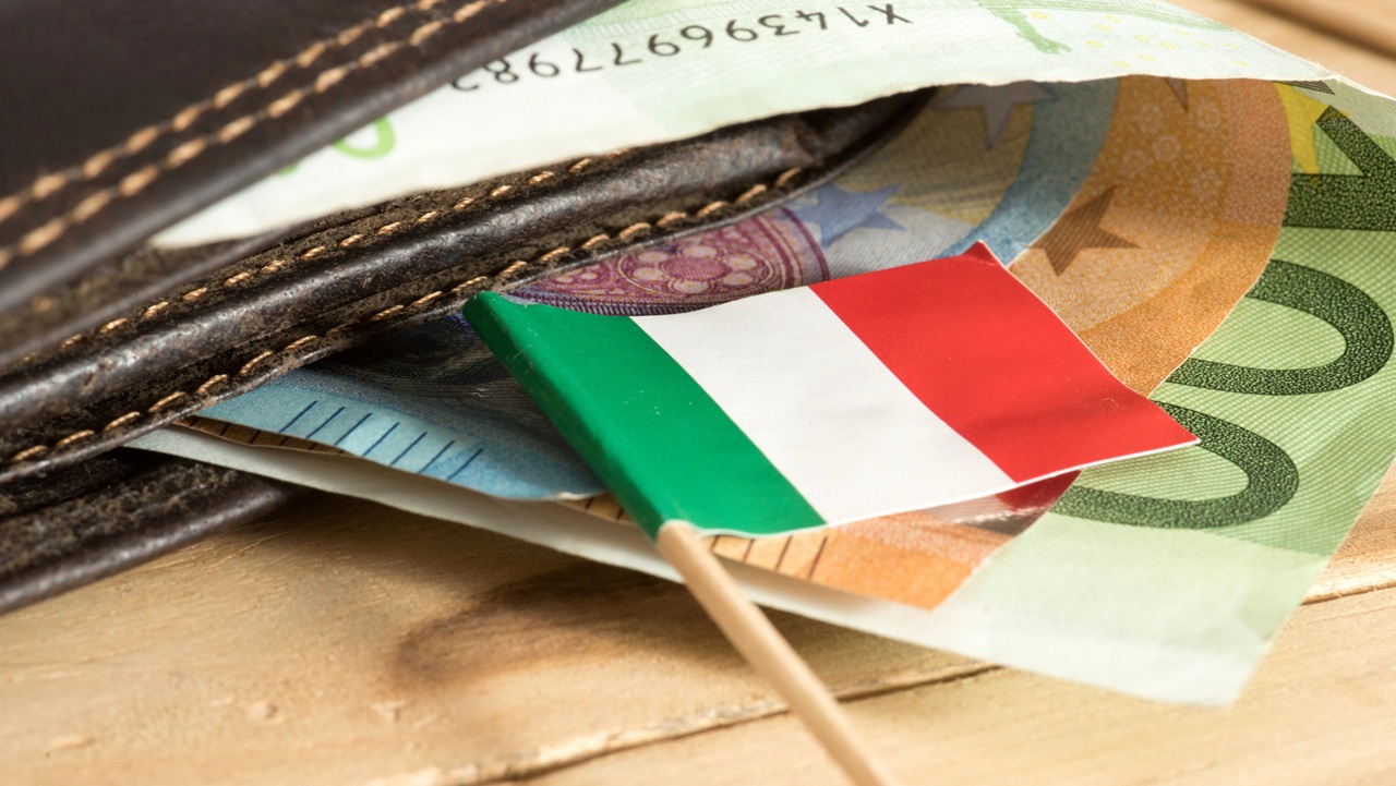 Una piccola bandiera italiana è appoggiata su banconote in euro che sporgono parzialmente da un portafoglio in pelle marrone appoggiato su una superficie di legno.