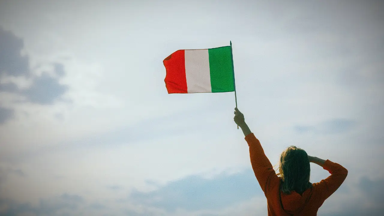 Una persona con i capelli lunghi tiene una bandiera italiana contro un cielo nuvoloso, dando le spalle alla telecamera.