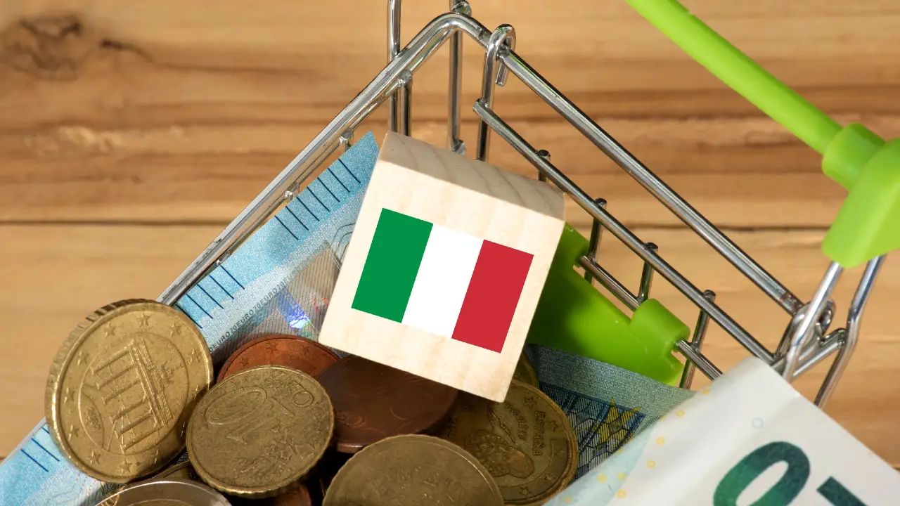 Un piccolo carrello della spesa pieno di monete e banconote in euro, con un blocco di legno con la bandiera italiana in cima.