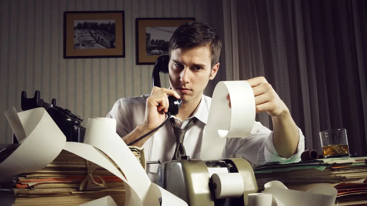 Un uomo stressato in camicia e cravatta è seduto a una scrivania disordinata, parlando al telefono mentre tiene in mano una lunga ricevuta. Documenti, cartelle, una calcolatrice e un drink sono sparsi intorno a lui in un ufficio scarsamente illuminato.
