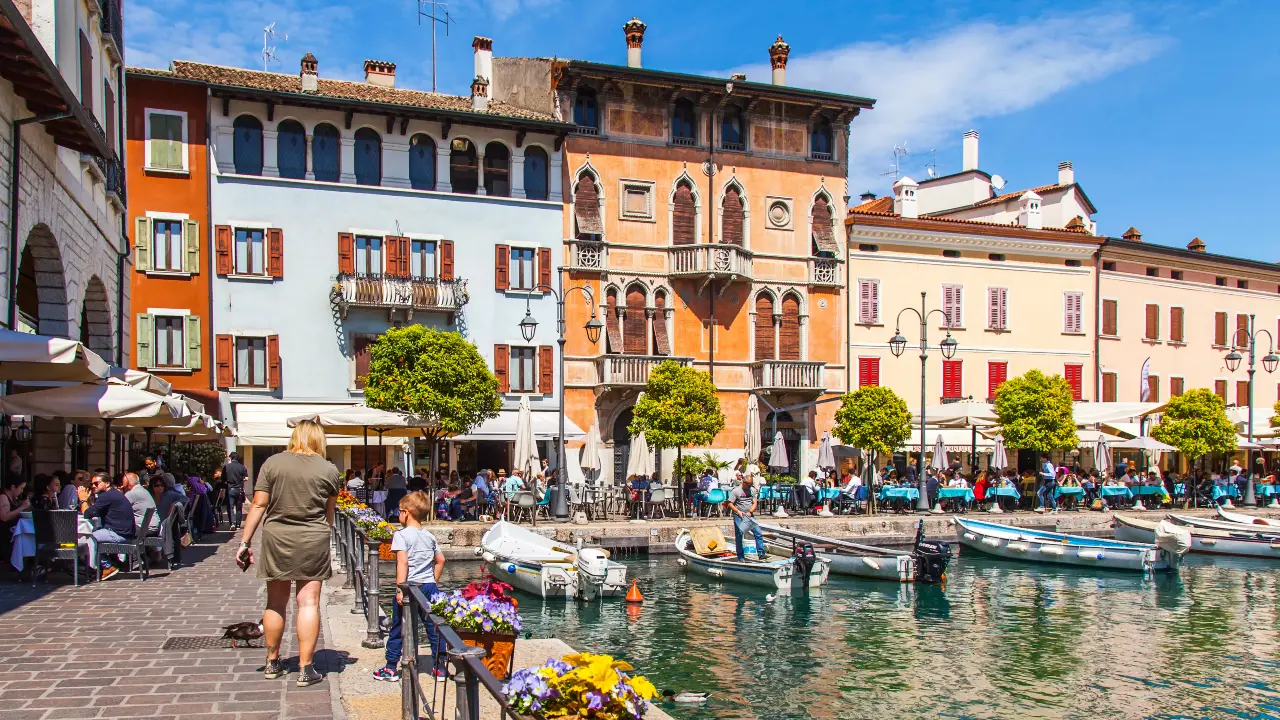 Immagine di un paese sul Lago di Garda con persone che camminano lungo un canale, piccole barche, edifici vivaci e caffè all'aperto, segno del fatto che sempre più investitori (anche) stranieri sono interessati al mercato immobiliare italiano.