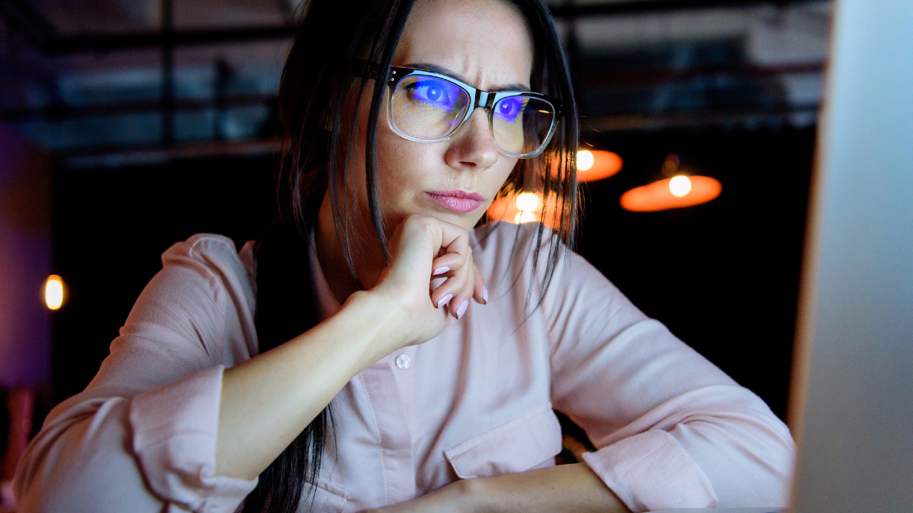 Una donna con gli occhiali e una camicetta rosa chiaro è seduta davanti a un computer, con il mento appoggiato sulla mano, mentre guarda pensierosa lo schermo, profondamente concentrata, forse impegnata in una seduta di consulenza. Luci calde illuminano lo sfondo.