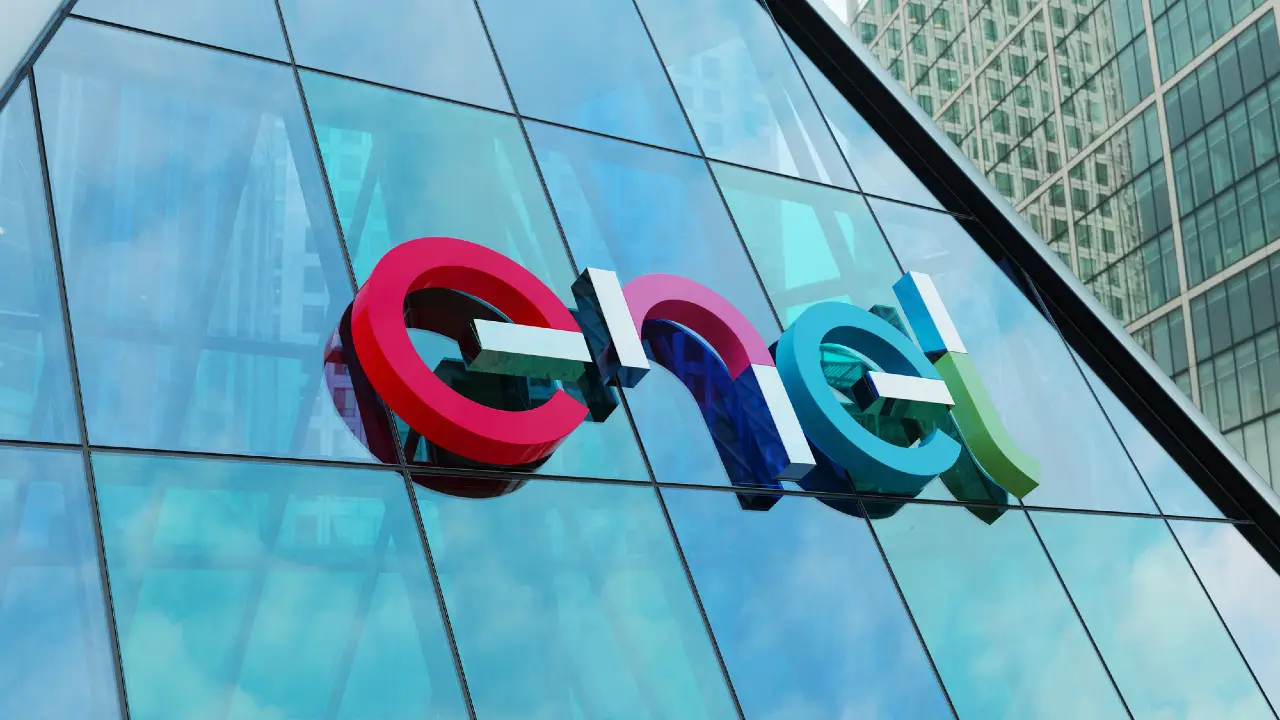 Il logo colorato dell'Enel è esposto sulla facciata in vetro di un moderno edificio per uffici, riflettendo il cielo e i grattacieli circostanti.