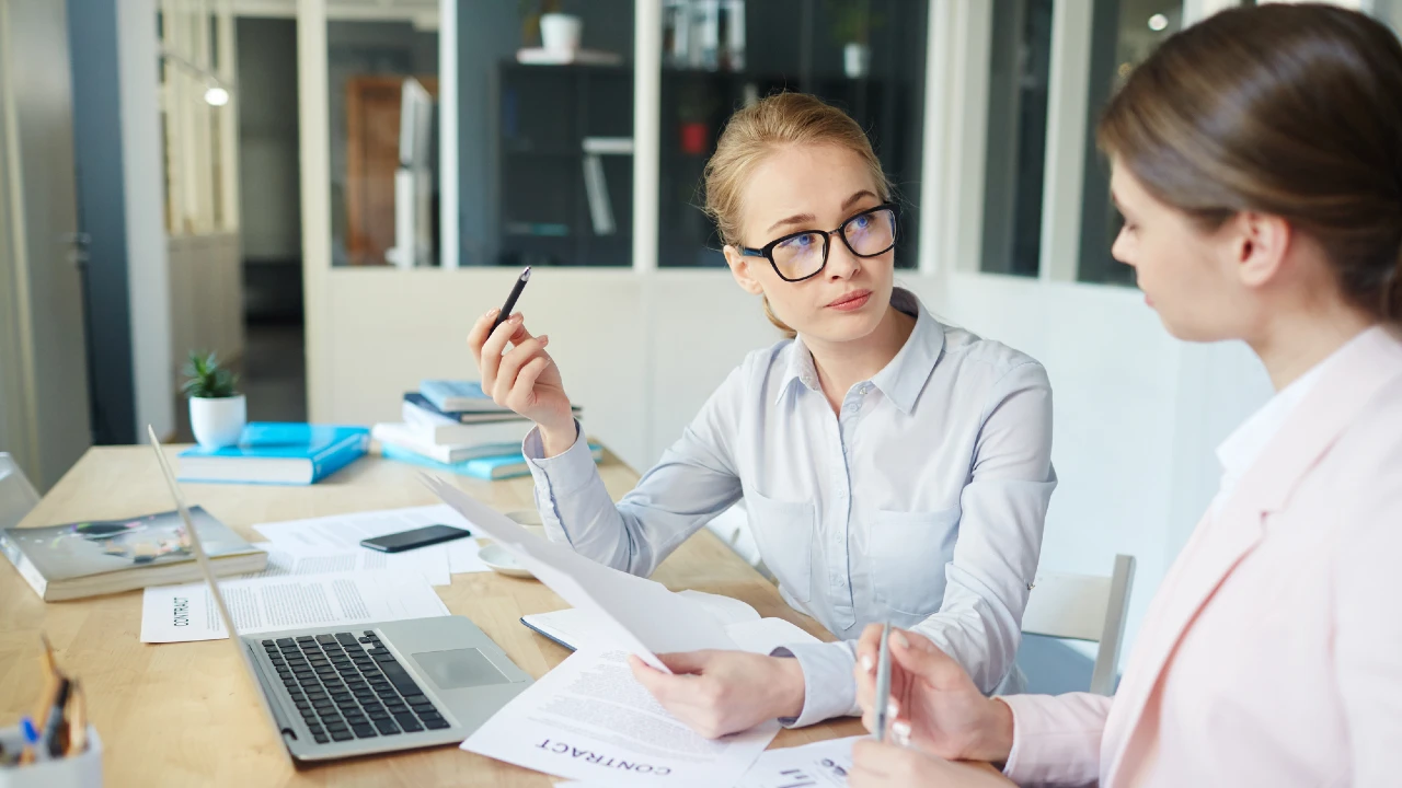 Due donne sono sedute a una scrivania in un ufficio, intente a discutere di documenti. Una donna, con gli occhiali, tiene in mano carta e penna mentre ascolta attentamente. Computer portatili, documenti e cartelle sono sparsi sulla scrivania.