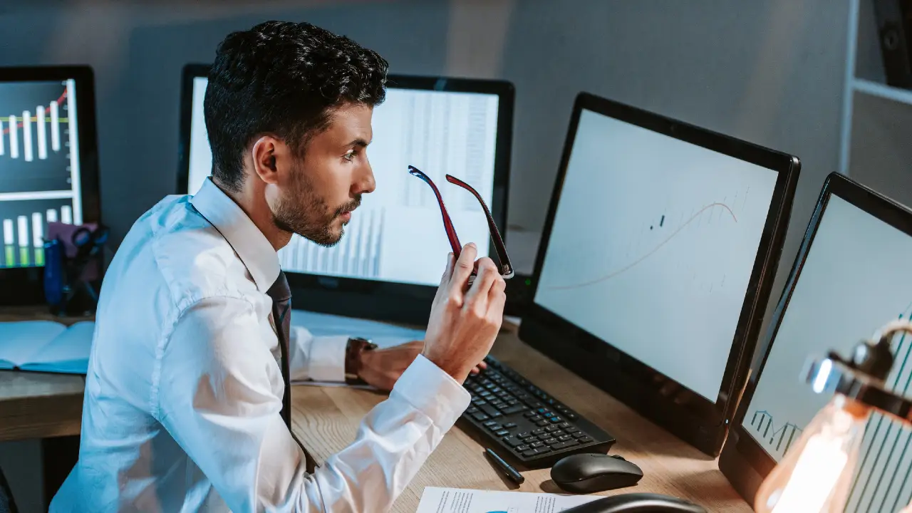 Un uomo in camicia bianca e cravatta tiene in mano gli occhiali mentre osserva attentamente il monitor di un computer che mostra un grafico a linee, mentre sulla sua scrivania ci sono altri due monitor che mostrano grafici di dati.