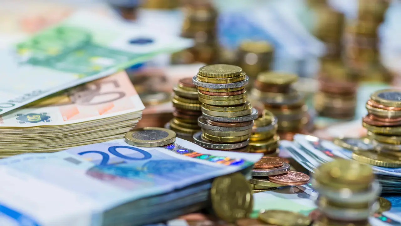 Pile di monete in euro e mazzette di banconote in euro di vari tagli sono distribuite su una superficie, a rappresentare ricchezza o transazioni finanziarie.