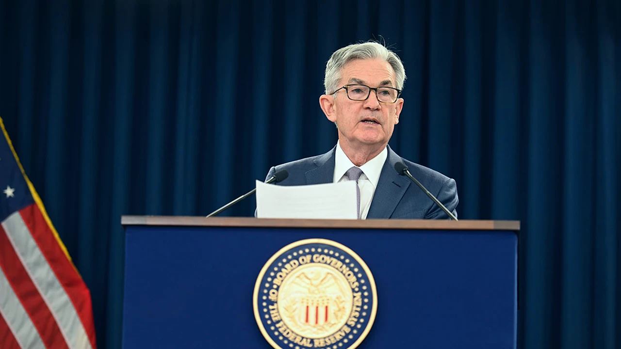 Un uomo in giacca e cravatta parla da un podio con il sigillo della Federal Reserve statunitense, tenendo in mano dei documenti. Sullo sfondo si vede una bandiera americana e dietro di lui si vedono delle tende scure.