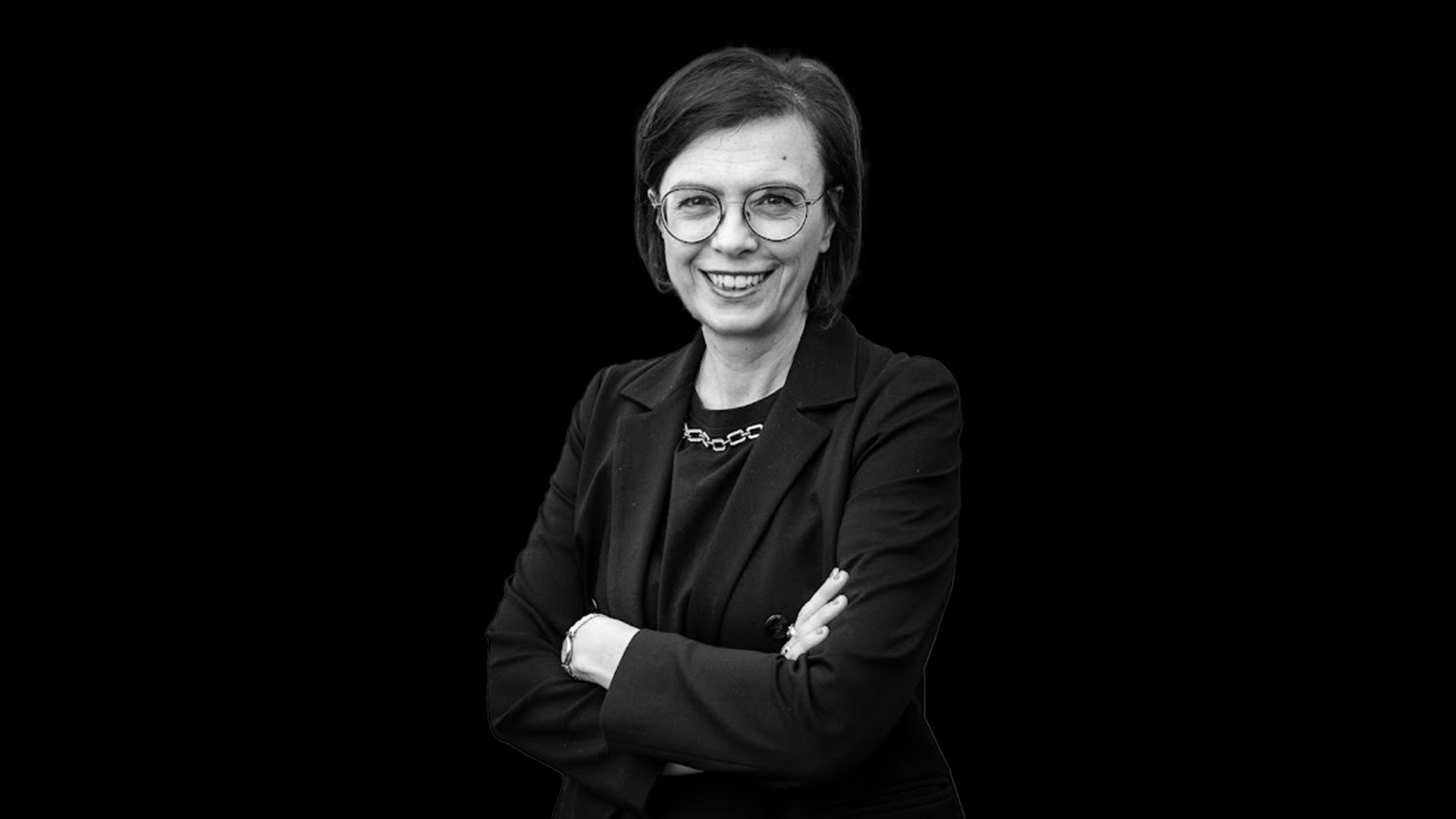 Una donna con i capelli corti e gli occhiali sorride, in piedi con le braccia incrociate. Indossa un blazer scuro, una collana a catena e un orologio: la sua posa sicura incarna lo stile senza tempo e la longevità della moda classica su uno sfondo nero uniforme.