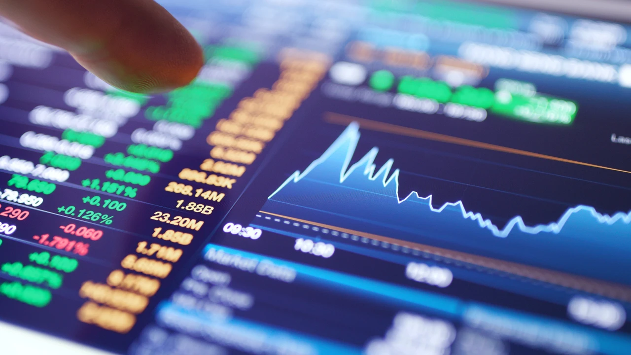 Primo piano dello schermo di un computer che mostra dati finanziari a colori, prezzi delle azioni in verde e arancione e un grafico blu che evidenzia le tendenze del mercato. Un dito punta verso lo schermo per indicare l’andamento dei mercati e la correlazione (o no) tra le varie asset class