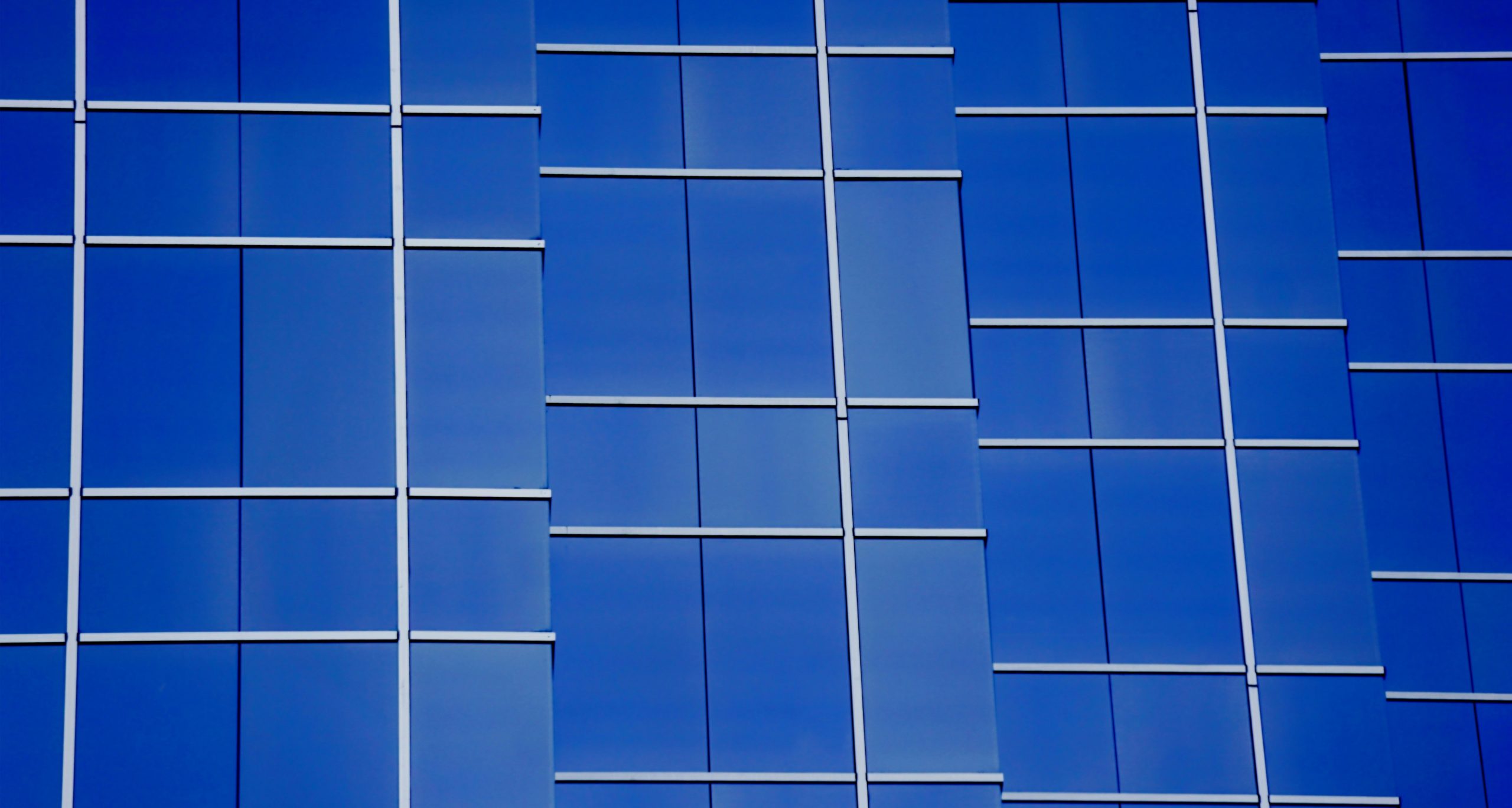 Primo piano della facciata di un edificio moderno con finestre in vetro blu riflettente e linee bianche a griglia che formano un motivo geometrico. Le finestre riflettono il cielo, creando un design elegante e astratto.