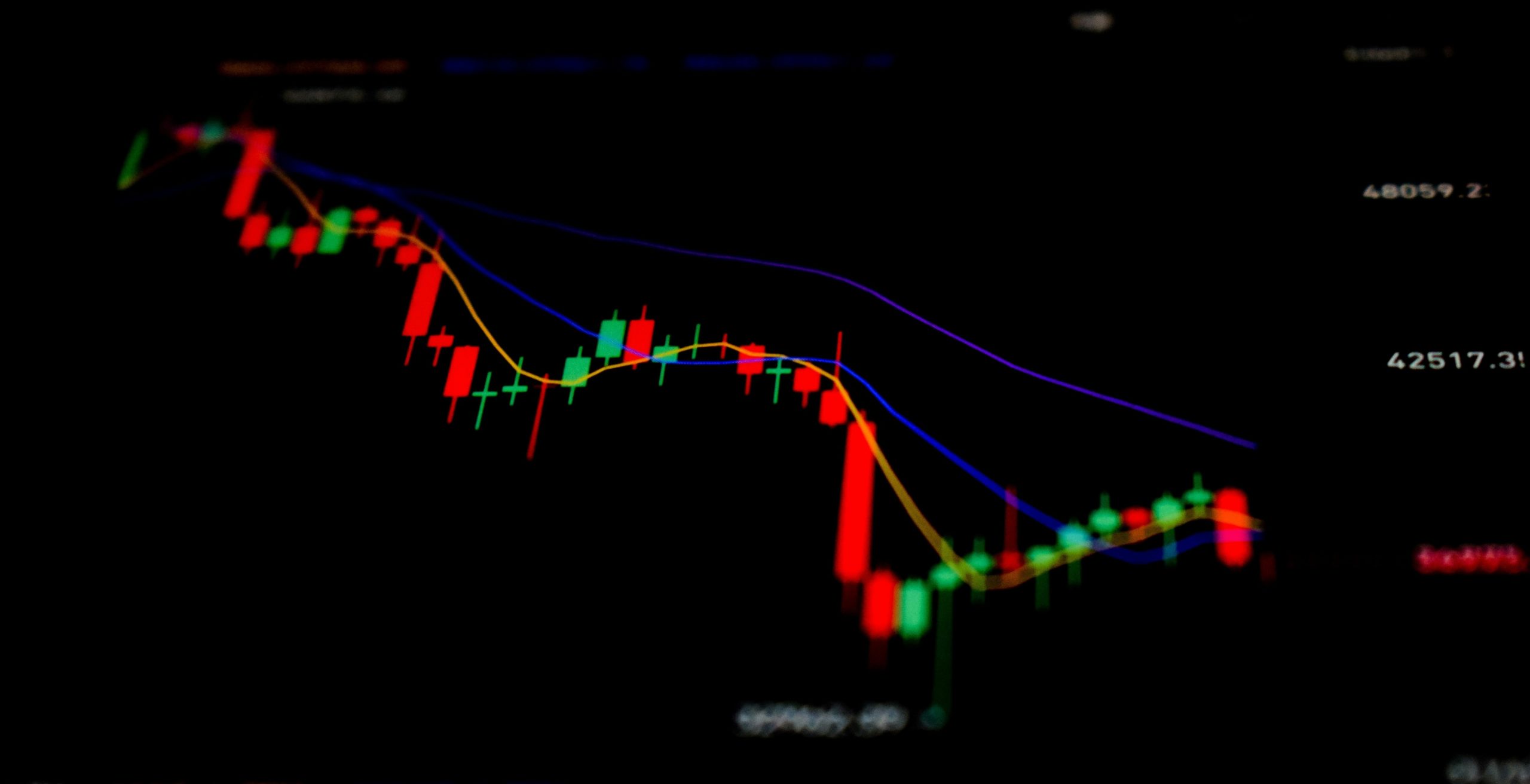 Un grafico a candele digitali che mostra un trend al ribasso, con barre rosse e verdi, due linee di media mobile colorate e valori dei prezzi su uno sfondo nero, che rappresentano l'attività del mercato finanziario.