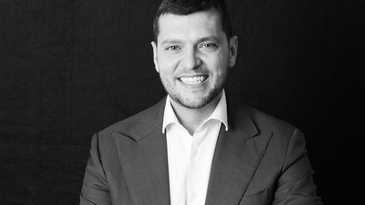 Un uomo in giacca e camicia bianca, rappresentante di Jupiter Asset Management, sorride alla telecamera su uno sfondo scuro e semplice. L'immagine è in bianco e nero.