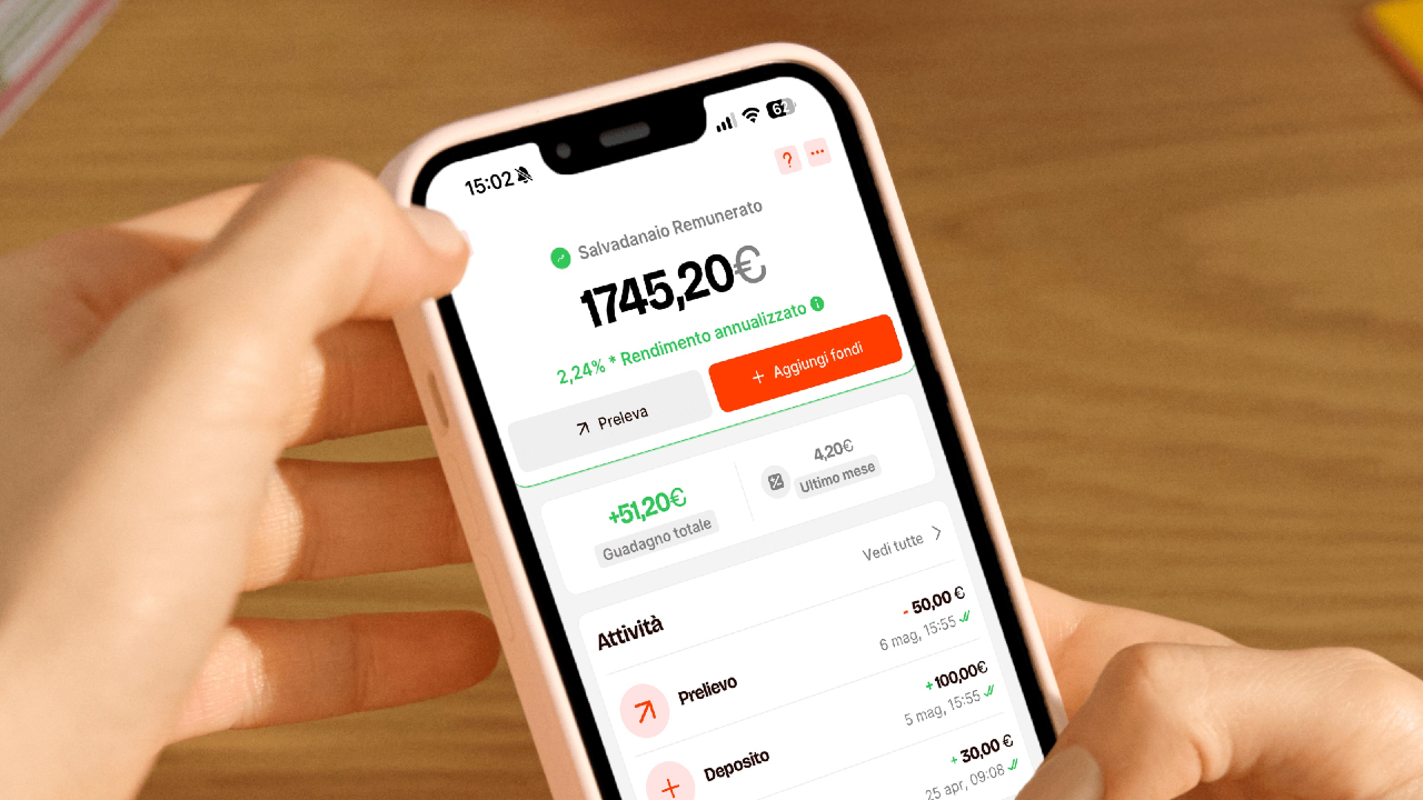 Una persona tiene in mano uno smartphone con un'app di risparmio in italiano, simile a Satispay, che mostra un saldo di 1745,20 euro e un pulsante verde con la scritta "Aggiungi fondi"; sullo sfondo c'è una superficie di legno.