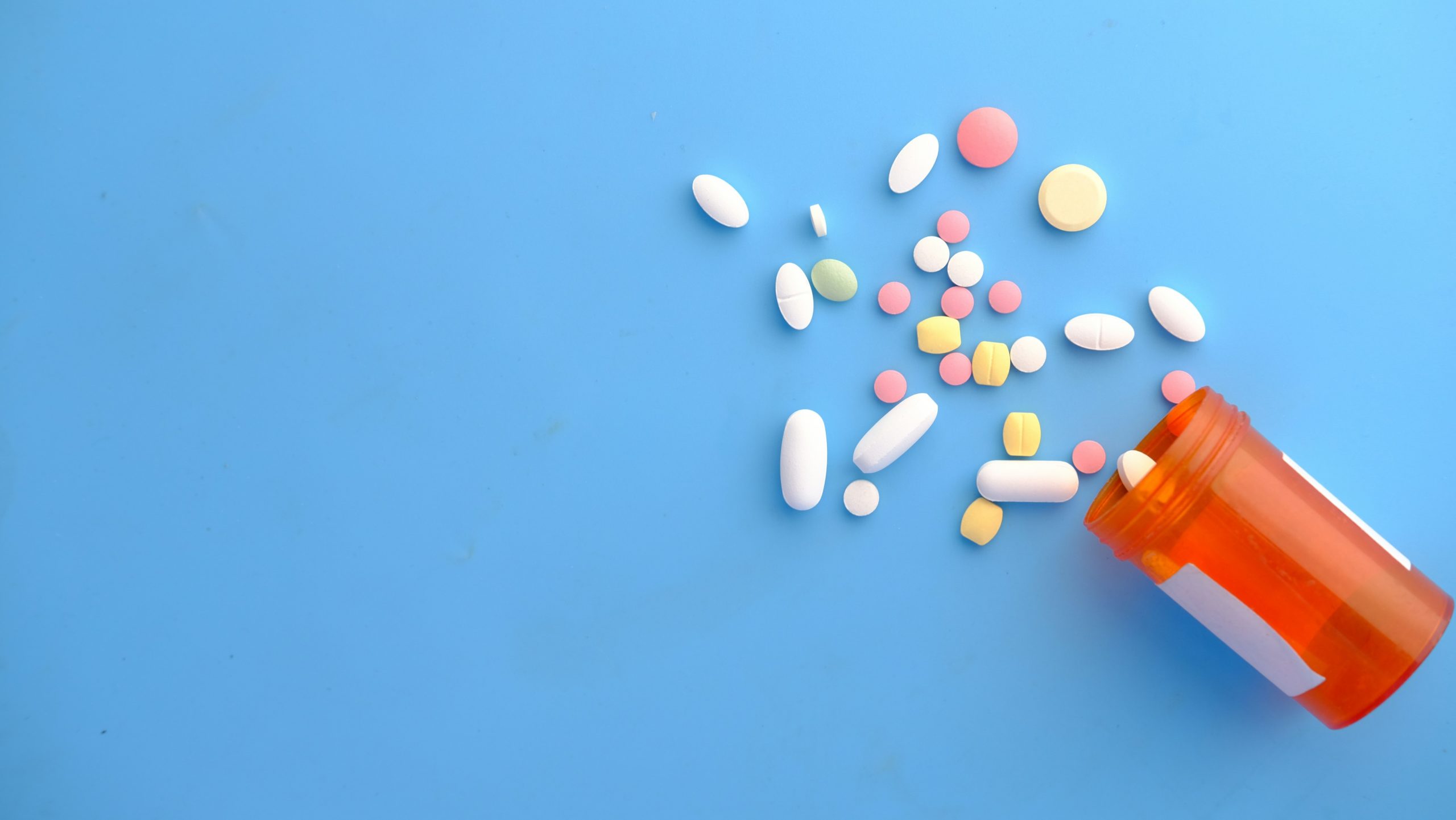Un flacone arancione di pillole da prescrizione si è ribaltato, rovesciando varie pillole e compresse colorate su una superficie blu.
