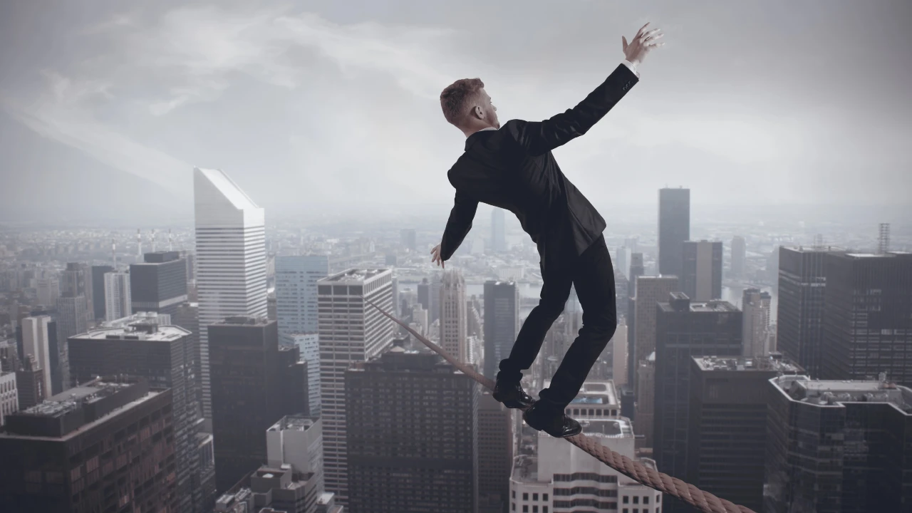Un uomo in giacca e cravatta cerca di stare in equilibrio su una corda tesa che sovrasta lo skyline di una città, con alti edifici e grattacieli sotto di lui, a simboleggiare i rischi che le imprese stanno affrontando a causa dei dazi Usa di Trump e le strategie da adottare per difendersi.