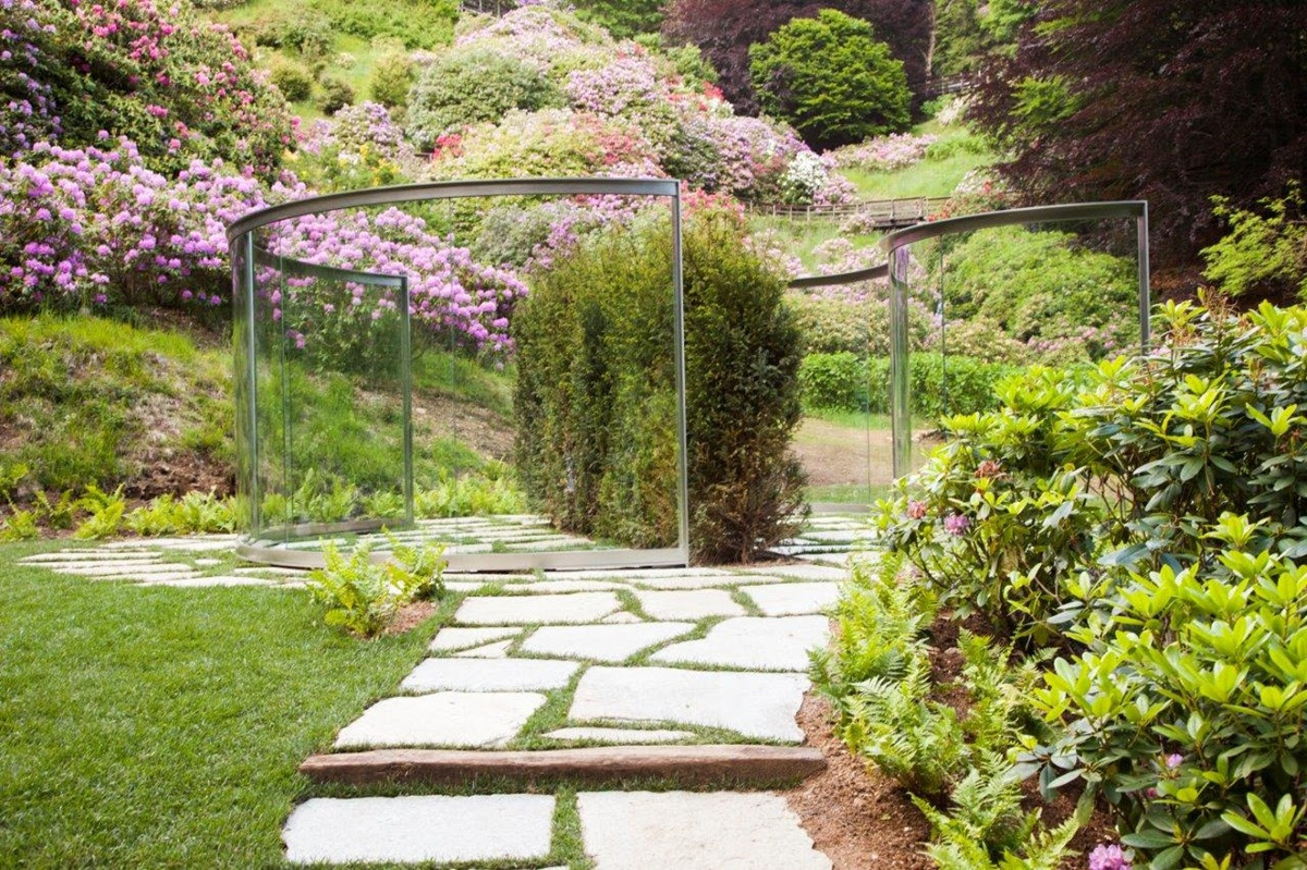 Pannelli di vetro curvi e alti arbusti formano un moderno labirinto da giardino su un sentiero in pietra, circondato da una vegetazione lussureggiante e da fiori rosa e viola in fiore sul pendio di una collina.