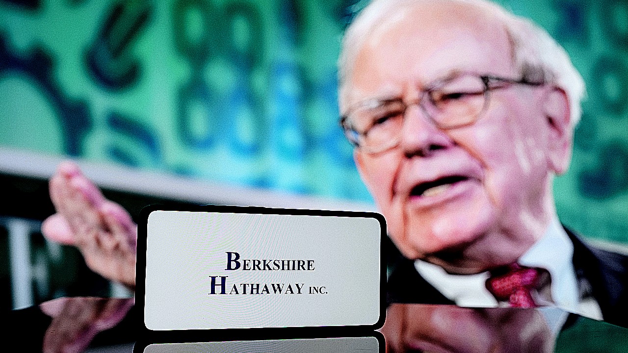 Un uomo anziano in giacca e cravatta gesticola mentre è seduto dietro un cartello con la scritta "Berkshire Hathaway Inc." Lo sfondo è sfocato, con toni blu e verdi.
