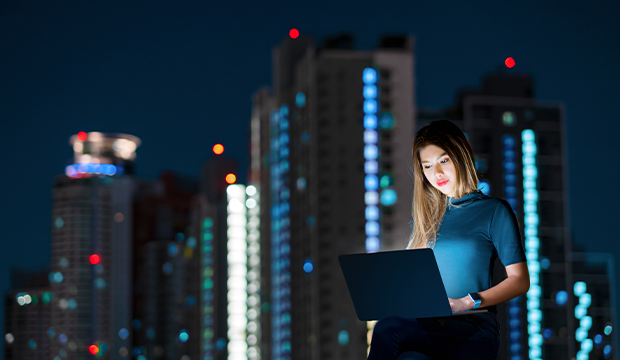 Una donna è seduta all'aperto di notte, illuminata dallo schermo del suo computer portatile, con alti edifici cittadini ben illuminati che si stagliano sullo sfondo.