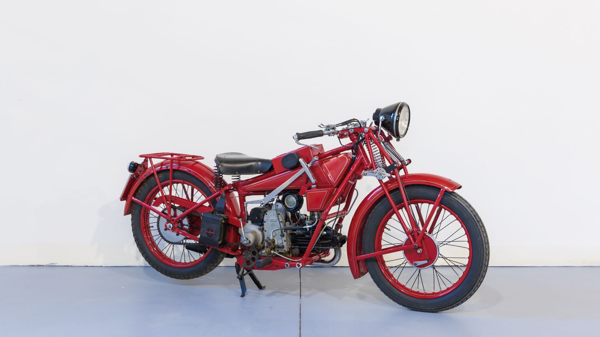 Una motocicletta rossa d'epoca con un grande faro, sella nera e motore a vista, esposta in un ambiente interno contro una semplice parete bianca e un pavimento azzurro.