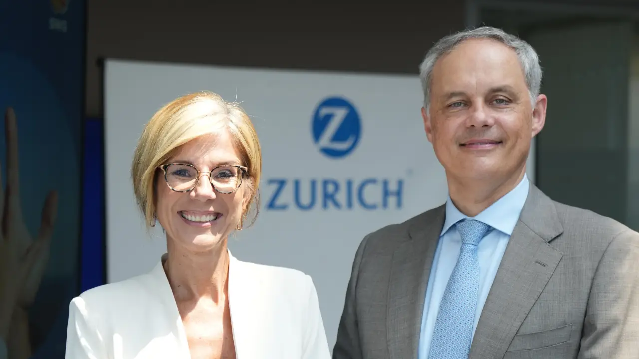 Due persone vestite in modo professionale, una donna e un uomo, sono in piedi sorridenti davanti a un cartello con il logo di Zurich Insurance. La donna ha i capelli corti e biondi e gli occhiali; l'uomo indossa un abito e una cravatta blu.