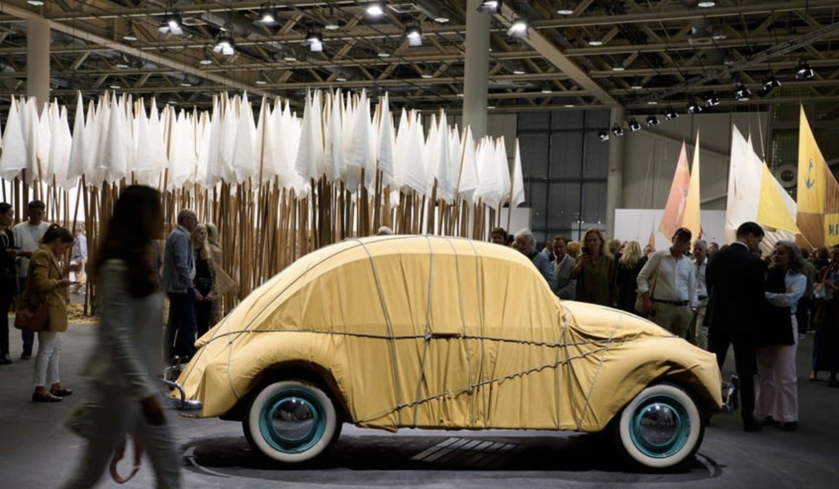 Un Maggiolino Volkswagen d'epoca rivestito di tessuto giallo è esposto all'interno, circondato da una folla. Sullo sfondo, alti pali con teli bianchi ricordano alberi astratti. La scena è ben illuminata e animata.