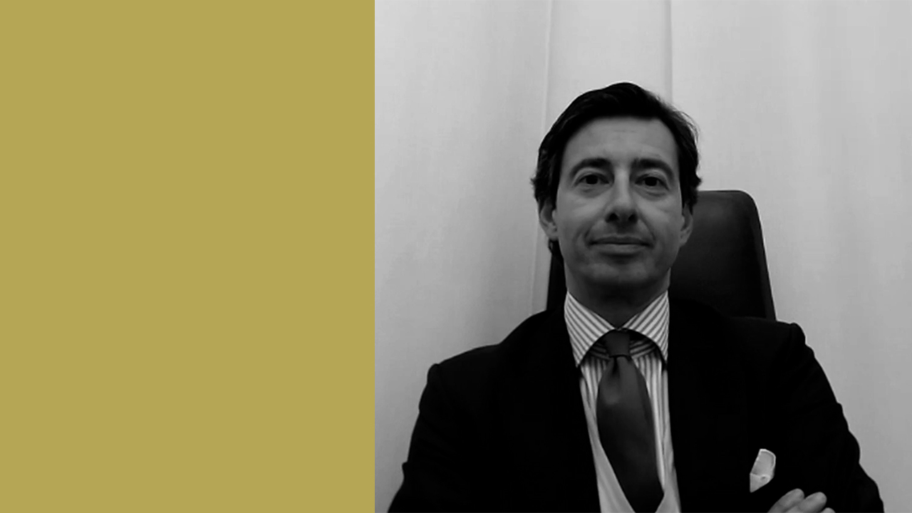 Un uomo in giacca e cravatta siede con le braccia incrociate, rivolto verso l'obiettivo su uno sfondo semplice con uno spacco verticale - oro a sinistra e bianco a destra - che cattura una serietà che non lascia spazio a nessuna clausola da baldracca.