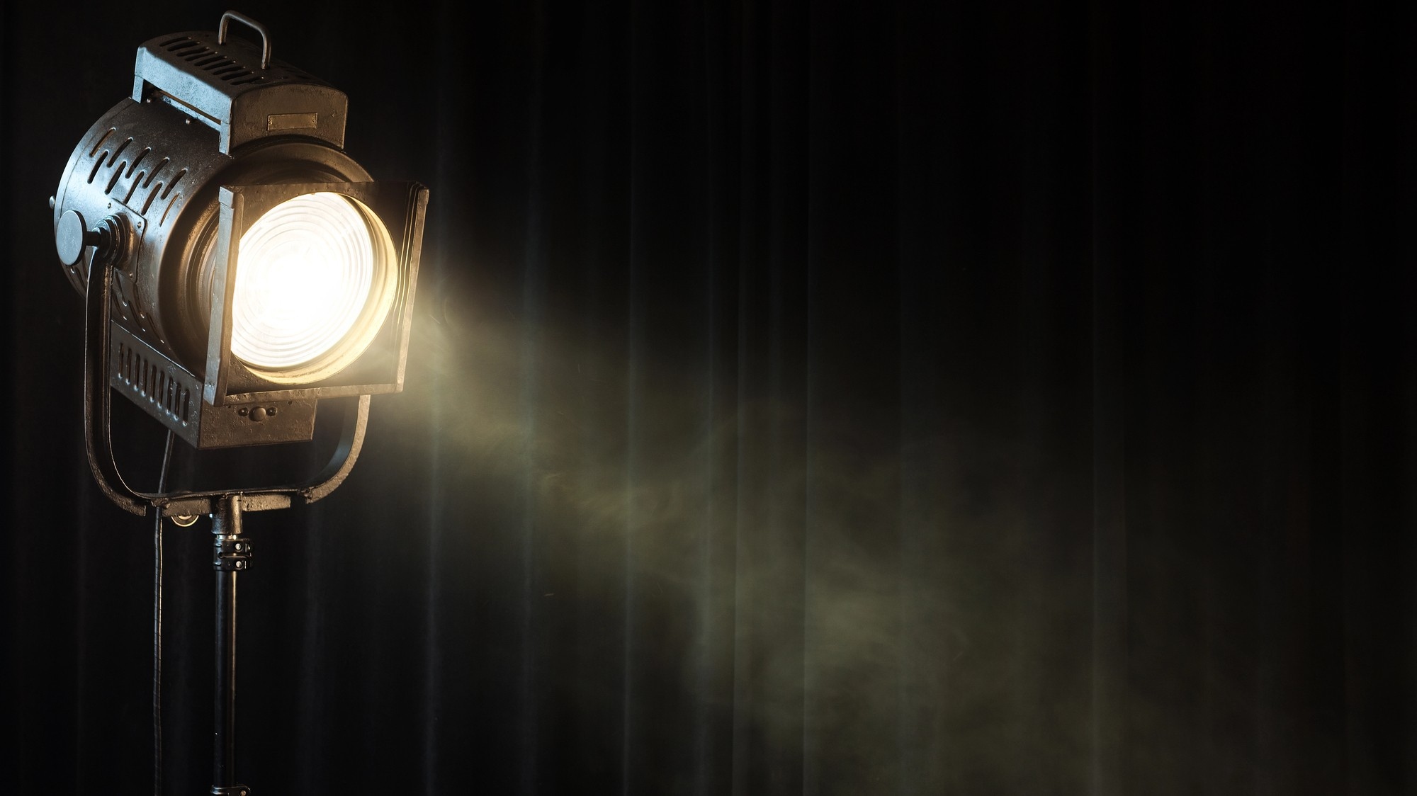 Un riflettore da palcoscenico d'epoca proietta un fascio di luce intensa nell'aria fumosa, sullo sfondo di una tenda nera e scura.