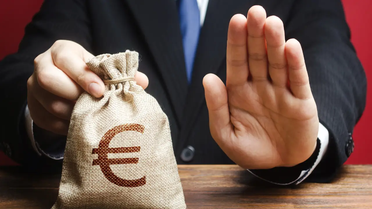 Una persona in giacca e cravatta fa cenno di fermarsi con una mano mentre con l'altra tiene un sacco di iuta con il simbolo dell'euro, suggerendo il rifiuto di denaro o di una transazione finanziaria.