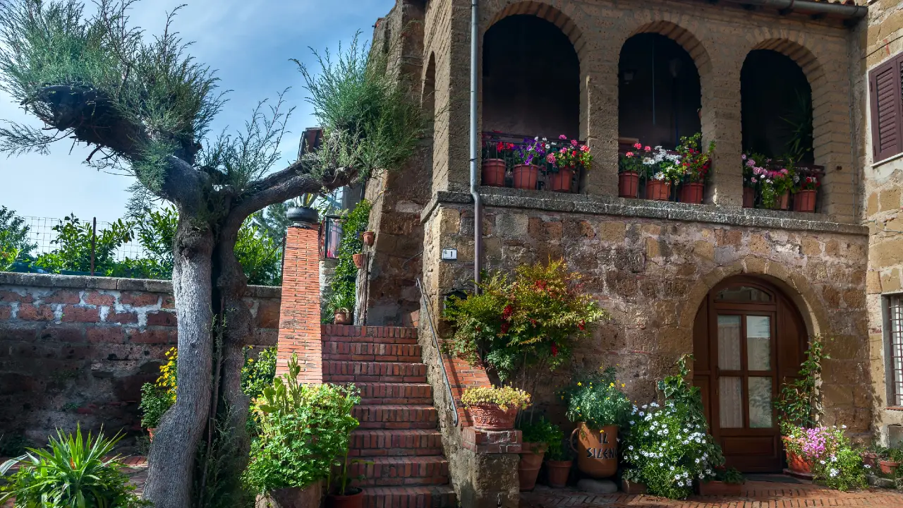 Una casa rustica in pietra con balcone ad arco, fiori in vaso e una porta di legno. I gradini in mattoni salgono accanto a un albero nodoso, circondato da piante verdi e fiori colorati sotto un cielo azzurro.