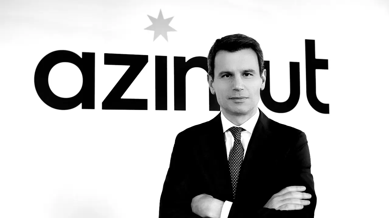 Un uomo in giacca e cravatta è in piedi con le braccia incrociate davanti a un muro su cui è stampata la parola "azimut" a caratteri cubitali. L'immagine è in bianco e nero.