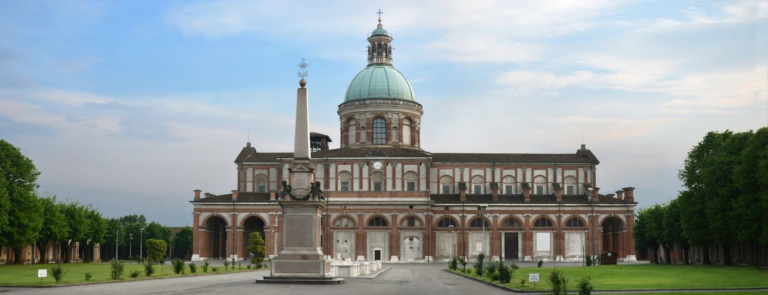 Una grandiosa basilica con una grande cupola centrale e colonne classiche si erge dietro un alto obelisco monumentale in un'ampia piazza verde fiancheggiata da alberi, sotto un cielo parzialmente nuvoloso.