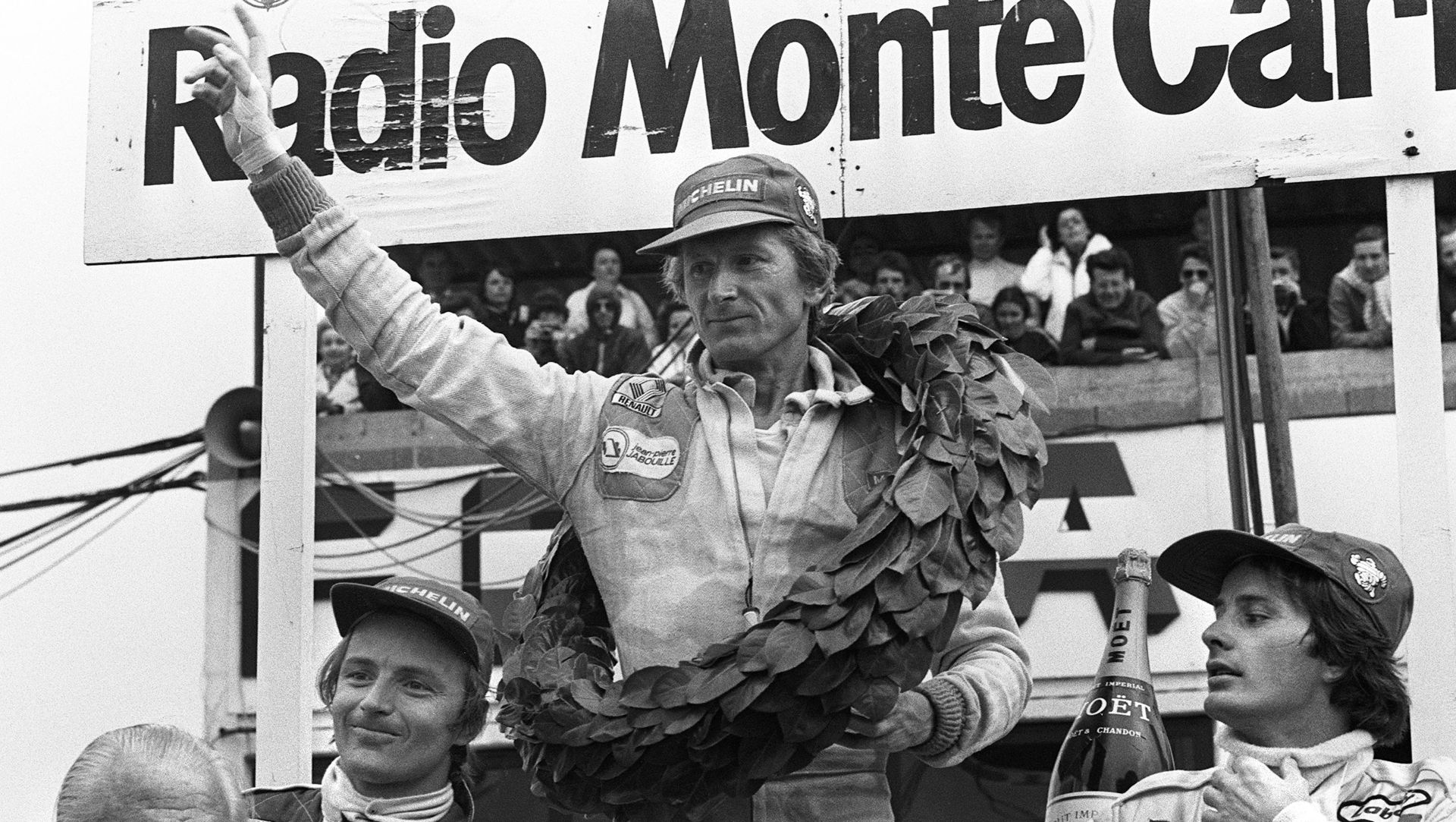 Un pilota automobilistico con una corona d'alloro alza un braccio in segno di vittoria su un podio, affiancato da altri due piloti. Una folla osserva da dietro un cartello "Radio Monte Carlo", celebrando il nuovo Rolex campione di Formula Uno.
