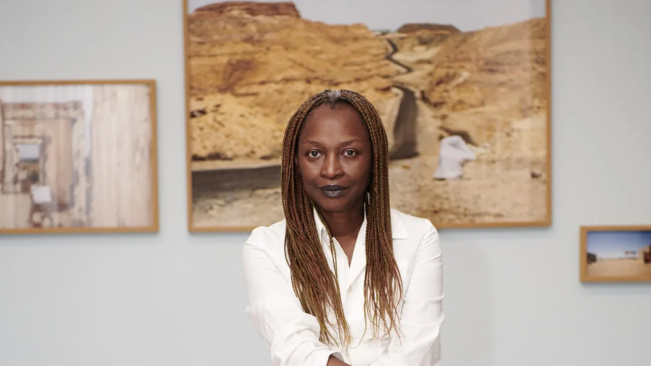 Una donna con lunghe trecce e rossetto scuro è in piedi davanti a fotografie incorniciate di paesaggi desertici, incarnando la forte presenza curatoriale di Koyo Kouoh, indossando una camicia bianca e guardando direttamente l'obiettivo.