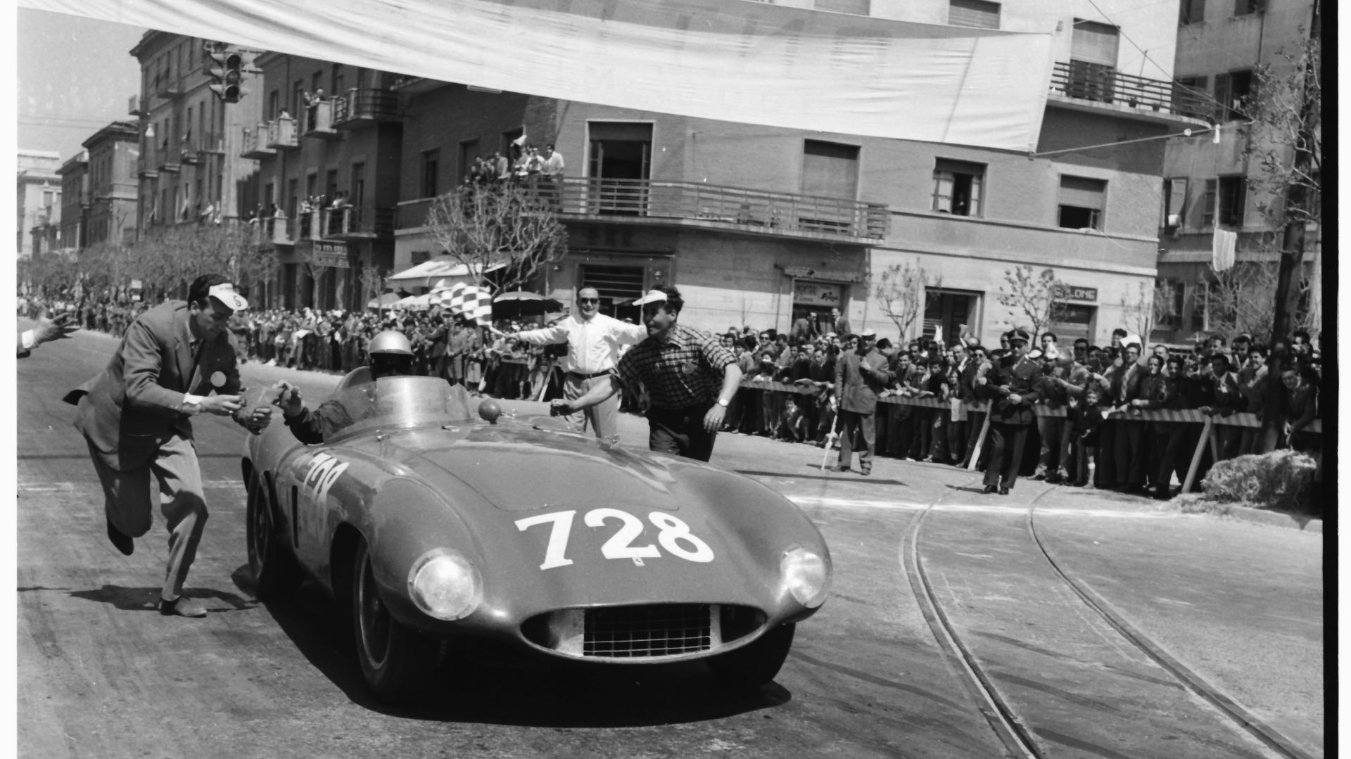 Un'auto da corsa d'epoca con il numero 728 taglia il traguardo in una strada cittadina durante la Mille Miglia 2025, sotto gli occhi di una folla. Una persona sventola una bandiera a scacchi mentre altri corrono accanto all'auto, festeggiando. La scena è in bianco e nero.
