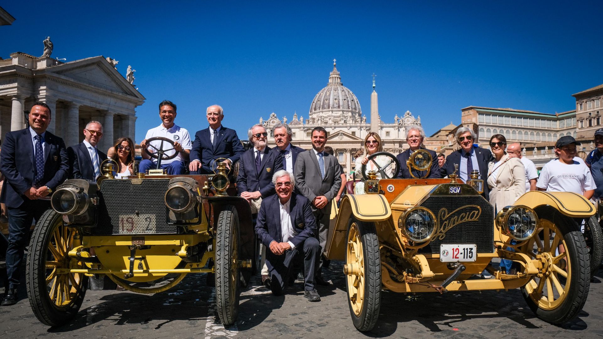 Un gruppo di persone posa dietro due auto gialle d'epoca in una piazza assolata, con la Basilica di San Pietro e un cielo azzurro sullo sfondo, suggerendo un evento di auto storiche giubileo in Vaticano.