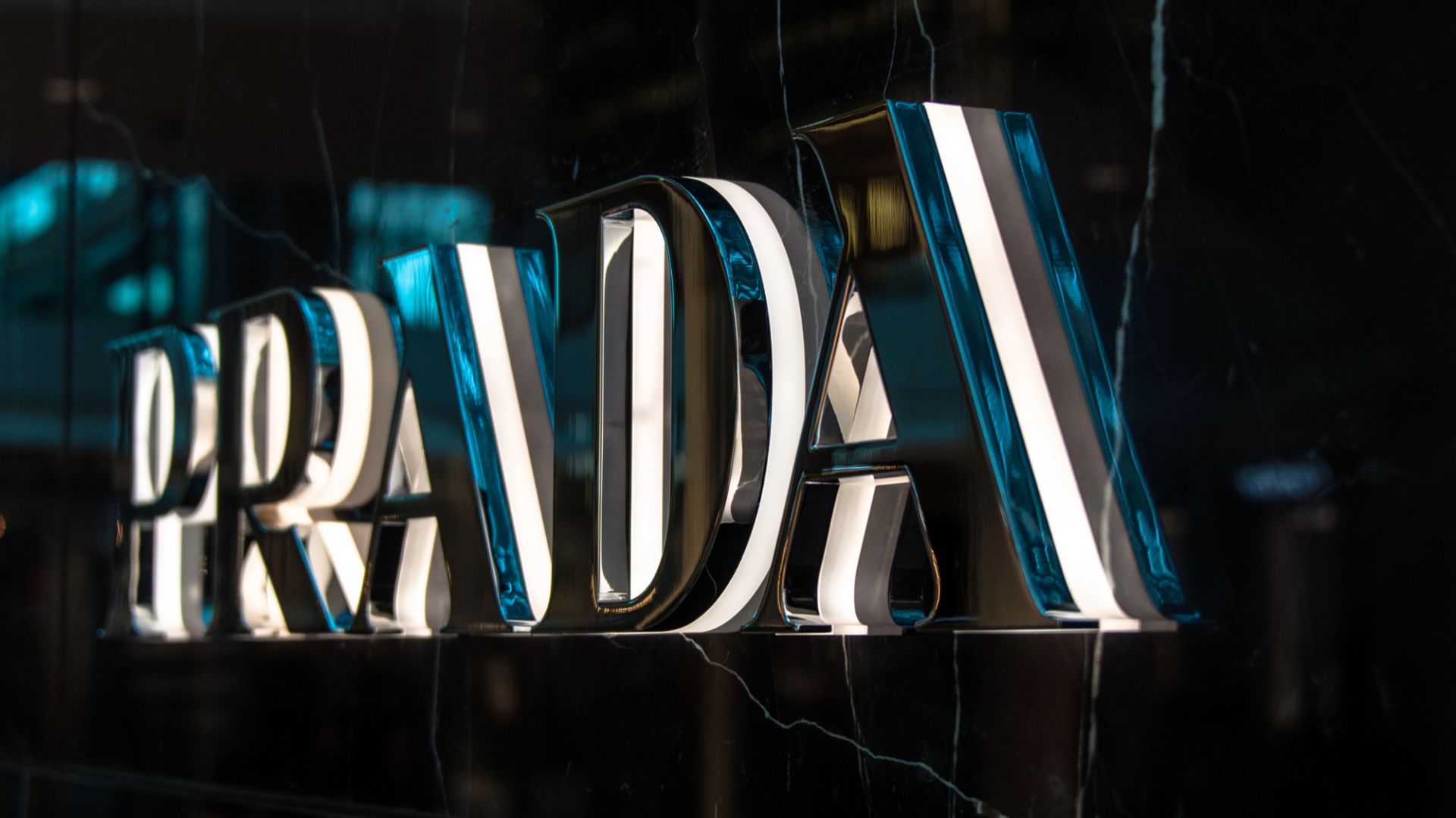 L'immagine mostra un primo piano di un'insegna metallica tridimensionale con il logo "PRADA" montata su una parete di marmo scuro e lucido, che riflette la luce e l'ambiente circostante, evocando l'eleganza dei nuovi cavalieri al lavoro.