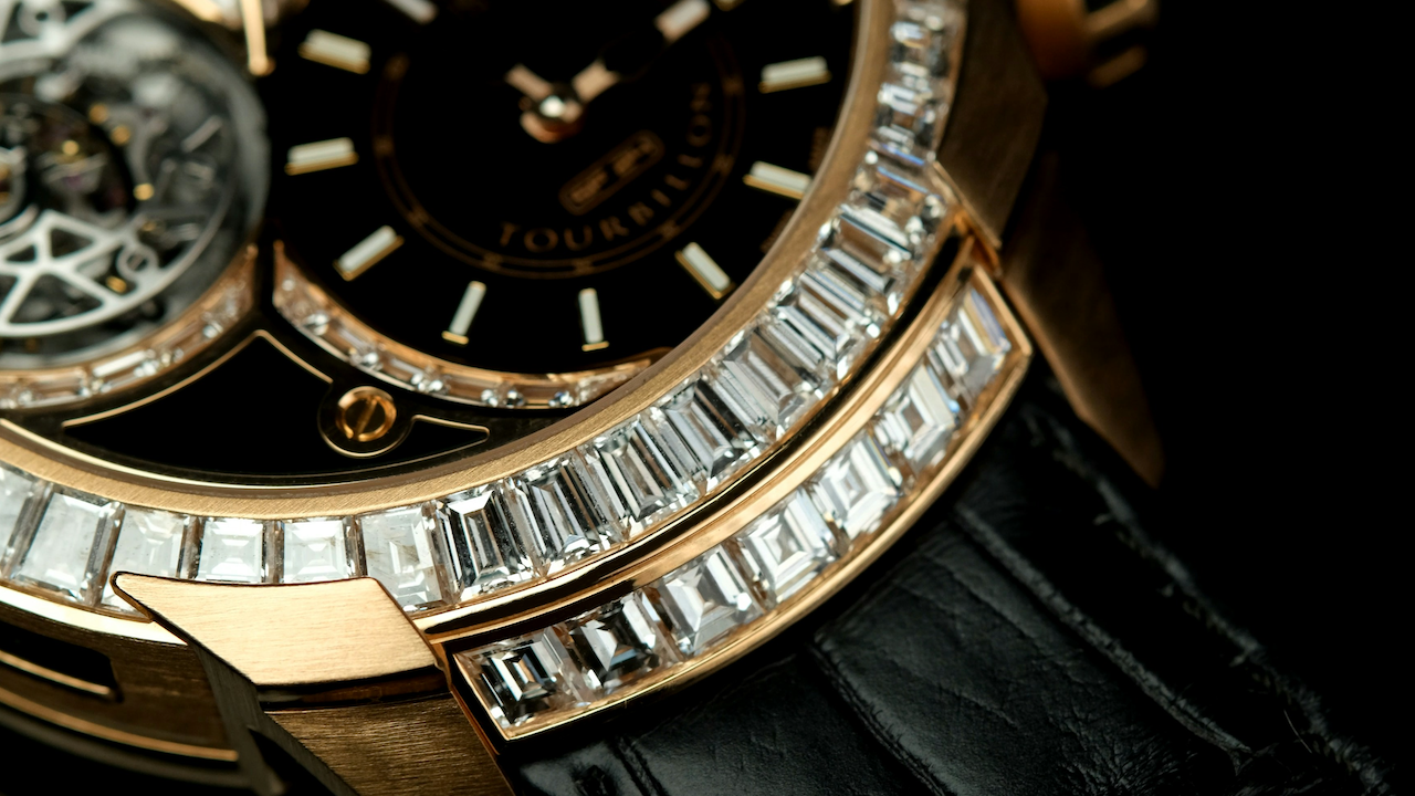 Primo piano di un orologio da polso di lusso con cassa in oro, quadrante nero, meccanismo tourbillon a vista e lunetta incastonata con diamanti rettangolari. L'orologio è dotato di cinturino in pelle nera.