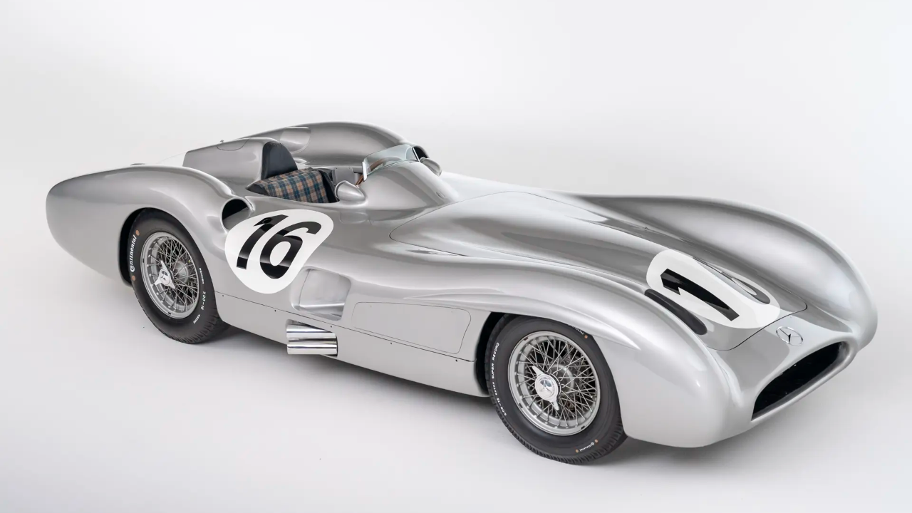 Un'auto da corsa d'epoca color argento con il numero 16 sulla fiancata e sul cofano, un abitacolo aperto con sedile a quadri, ruote a raggi metallici e un design aerodinamico della carrozzeria, fotografata in uno studio bianco.