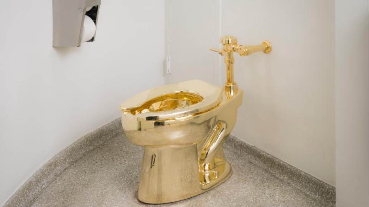 Una toilette color oro lucido è installata nell'angolo di un bagno con pareti chiare e pavimento grigio a chiazze; un distributore di carta igienica è montato sulla parete vicina.
