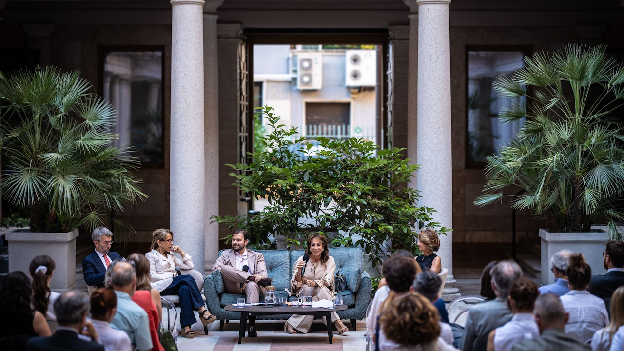 Un gruppo di cinque persone siede su un divano e delle sedie, parlando a un pubblico in un cortile con colonne e piante in vaso, durante un evento diurno.
