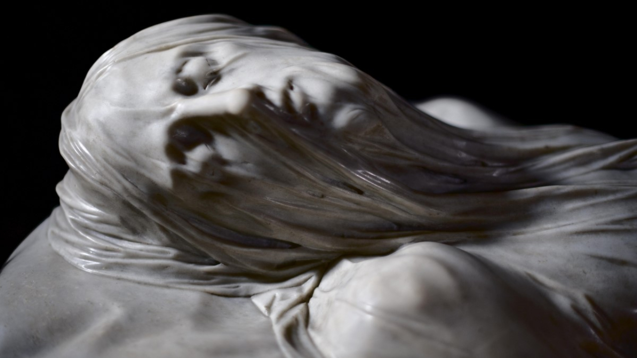 Primo piano di una scultura in marmo che raffigura una figura con un velo trasparente e fluente sul volto, che rivela i delicati tratti del viso sotto la pietra traslucida. Lo sfondo è scuro e mette in risalto gli intricati dettagli della scultura.
